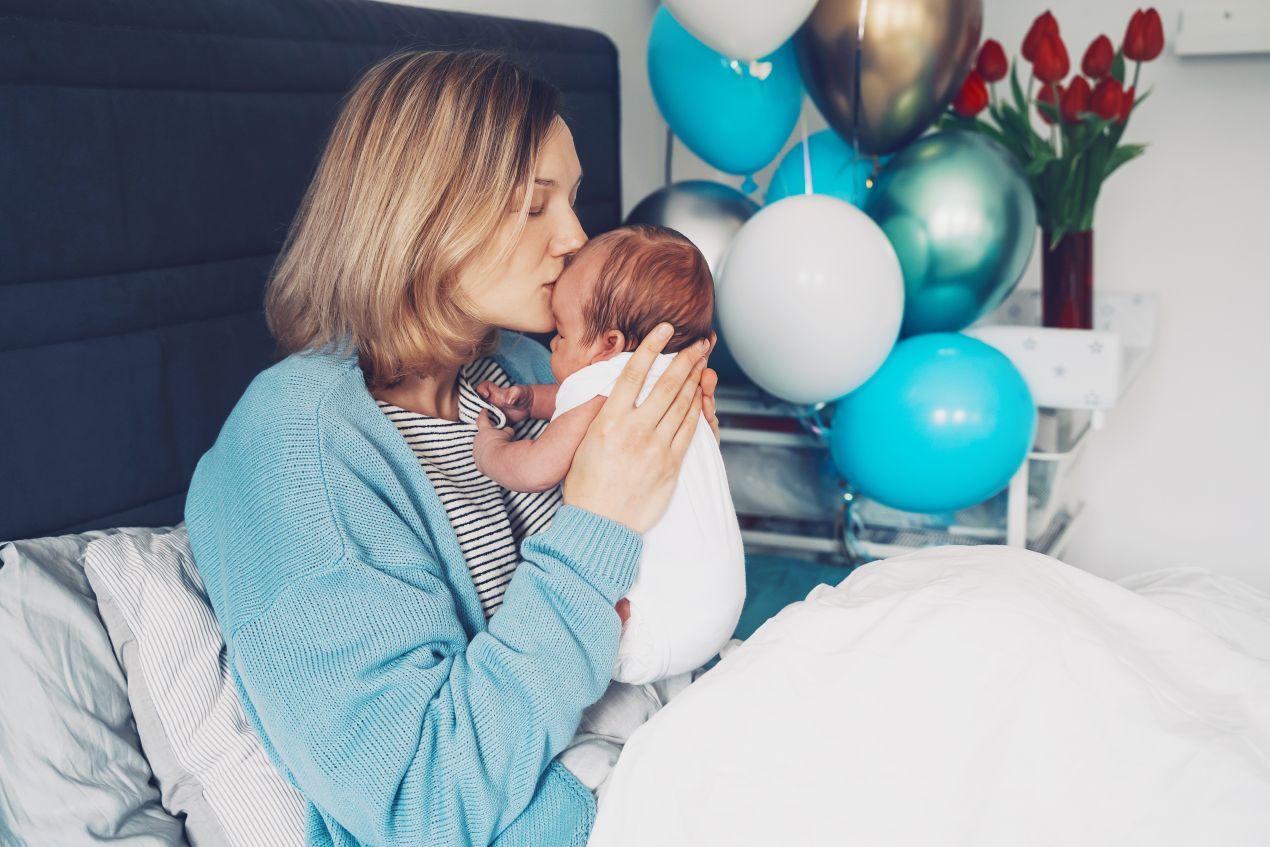Geschenk zur Geburt fuer Mutter mit Neugeborenem, blauen Luftballons und frischen Blumen